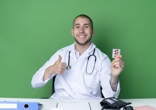 医生面带微笑的年轻男医生穿着医用长袍 听诊器坐在办公桌旁 手里拿着一包胶囊 在绿色的墙上孤立地竖起大拇指工具胶囊表情