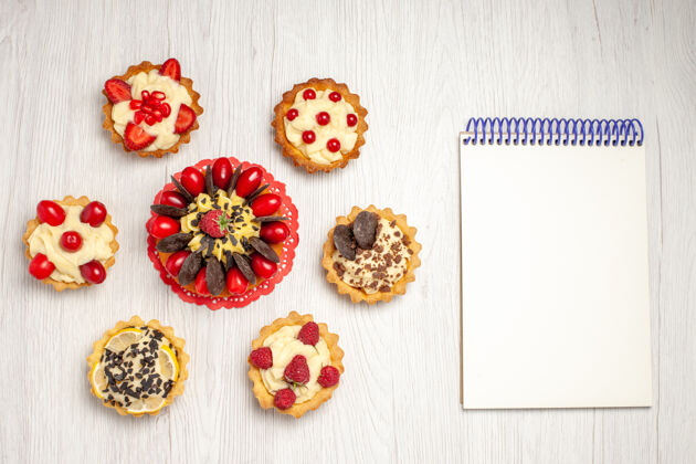 食品俯瞰红色椭圆形蕾丝doily馅饼上的浆果蛋糕和白色木质地面上的笔记本粉色形状笔记本