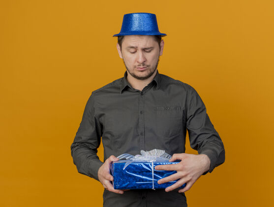 年轻困惑的年轻人戴着蓝色的聚会帽 拿着礼物盒看着孤立的橙色帽子盒子蓝色