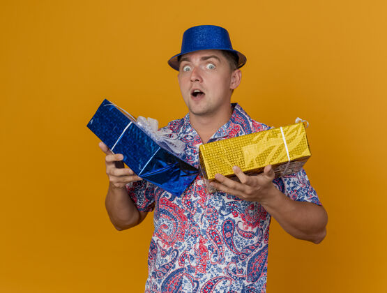 惊喜惊讶的年轻人戴着蓝色帽子拿着礼品盒孤立的橙色穿蓝色年轻