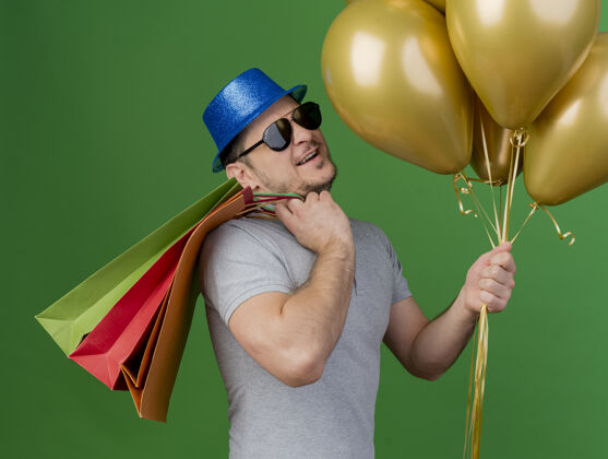 包微笑的年轻人戴着派对帽和眼镜 肩上挎着礼品袋 手里拿着绿色的气球礼物肩膀眼镜