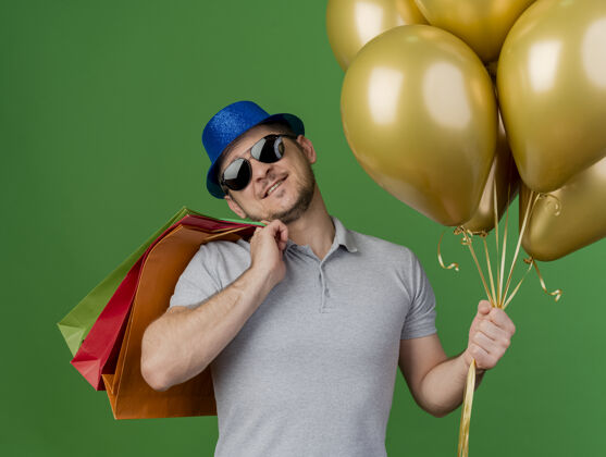 肩微笑的歪着头的年轻人戴着聚会帽和眼镜把礼物袋放在肩膀上 拿着气球孤立在绿色聚会穿眼镜