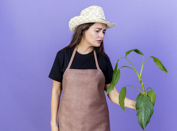 悲伤悲伤美丽的园丁女孩穿着制服 戴着园艺帽 抱着一株孤零零地看着蓝色的植物帽子举行穿