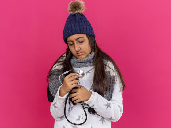 围巾年轻的生病的女孩戴着冬天的帽子戴着围巾拿着听诊器看着羽绒服隔离在粉红色穿抱听诊器