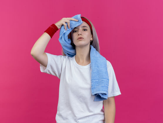 自信自信的年轻运动女性戴着背带 戴着头带和腕带 用隔离在粉色墙上的毛巾托住并擦拭额头人女孩抱着