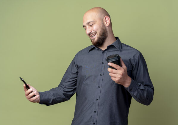 橄榄微笑着的年轻秃头呼叫中心男子手持手机 看着另一只手拿着塑料咖啡杯孤立在橄榄绿的墙上握着绿手机