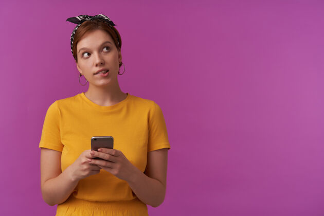 头巾自然妆容的女人 穿着黄色t恤 黑色大手帕 手拿电话 情绪化的脸 在紫色的墙上看着一边衬衫衣服看一边