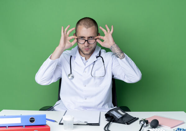姿势面容严肃的年轻男医生穿着医用长袍和听诊器坐在办公桌旁 带着工作工具 戴着眼镜 看着隔离在绿色墙上的前面桌子严肃工具