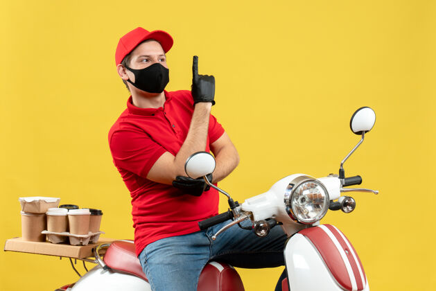 摩托车顶视图自信的年轻人穿着红色上衣 戴着帽子手套 戴着医用面罩 坐在踏板车上传递命令 黄色背景上有一个男人年轻人医学