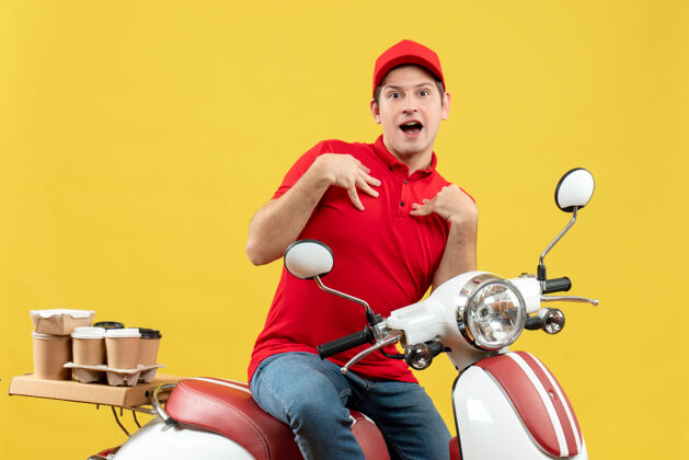 年轻人上图是困惑的年轻人穿着红色上衣 戴着帽子 坐在踏板车上 指着黄色的背景 传递着命令帽子指着命令