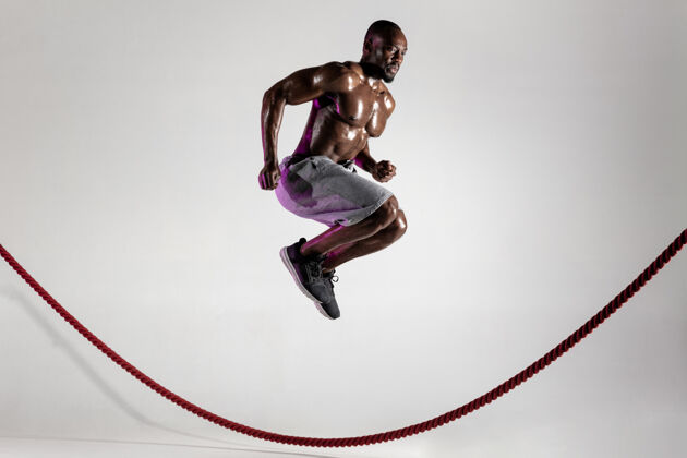 手第一个年轻的非洲裔美国人在灰色工作室背景下的健美训练穿着运动服的肌肉男模特跳过战绳运动的概念 健美 健康的生活方式手臂绳子举重