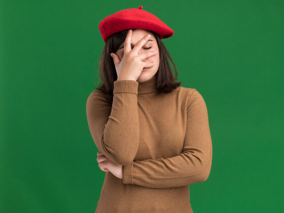 绿失望的年轻漂亮白人女孩戴着贝雷帽把手放在绿色的脸上贝雷帽漂亮帽子