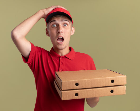 橄榄焦急的年轻金发送货员把手放在头上 拿着橄榄绿的比萨饼盒盒子焦虑金发