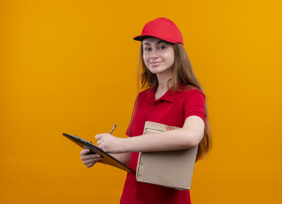 女孩高兴的年轻送货女孩在红色制服举行的盒子和书写在剪贴板上孤立的橙色空间红色剪贴板写字