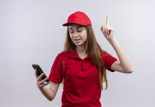 制服令人印象深刻的年轻送货女孩在红色制服举行 并在孤立的白色空间抬起手指看手机电话送货拿着