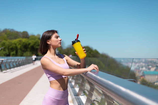 休闲年轻女子穿着合身的运动服 在炎热阳光明媚的早晨 在桥上喝着一瓶水 运动后口渴又累身体震动女人