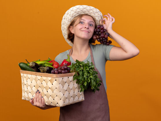 蔬菜高兴的年轻斯拉夫女园丁戴着园艺帽 手里拿着菜篮子和橘子上的葡萄葡萄斯拉夫人幼的