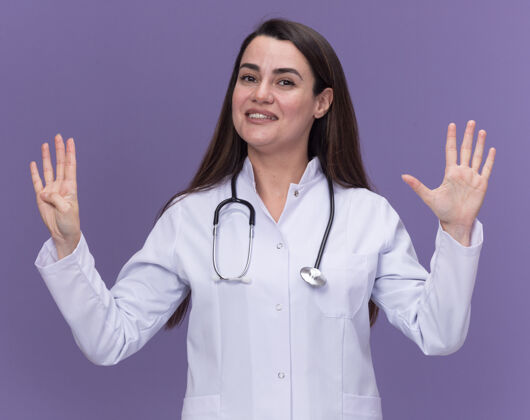 手指面带微笑的年轻女医生穿着医用长袍 手持听诊器 用九根手指在紫色上做手势年轻九长袍