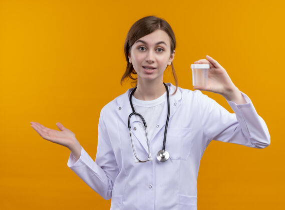 女性年轻的女医生穿着医用长袍 手持听诊器 拿着烧杯 在孤立的橙色空间里空手而出医学穿橙色