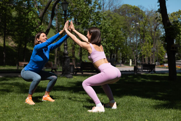 瑜伽阳光明媚的日子里 两个穿着运动服的漂亮女人在公园的草地上锻炼 蹲着支撑着彼此快乐的情绪放松健身锻炼