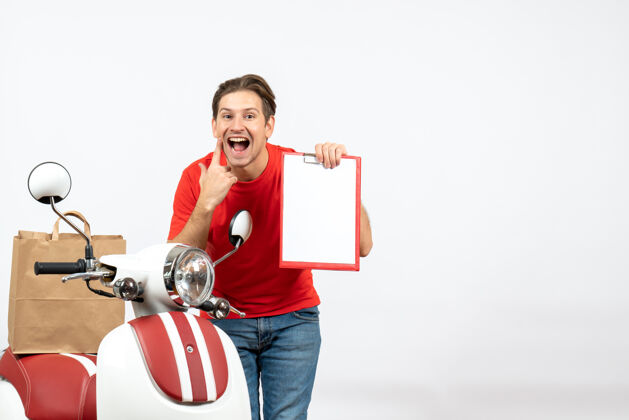 滑板车身穿红色制服的年轻快乐送货员站在滑板车旁 手里拿着白墙上的文件站着靠近文件