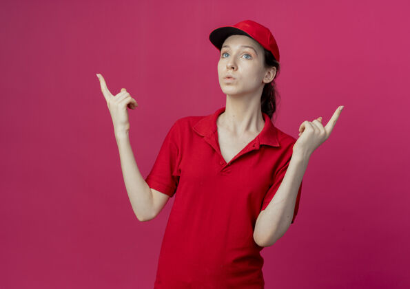 制服令人印象深刻的年轻漂亮的送货女孩在红色制服和帽子看直 并指出在深红色背景与复制空间孤立向上女孩帽子