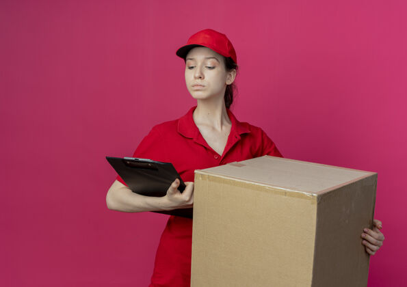 红色自信的年轻漂亮的送货女孩 穿着红色制服 戴着帽子 手里拿着纸盒和剪贴板 看着被隔离在深红色背景上的剪贴板 还有复印空间盒子背景剪贴板