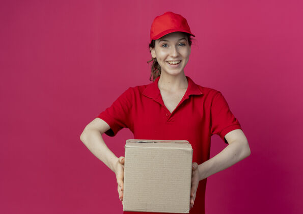 漂亮快乐的年轻漂亮的送货女孩在红色制服和帽子举行纸箱在深红色背景与复制空间隔离持有帽子年轻