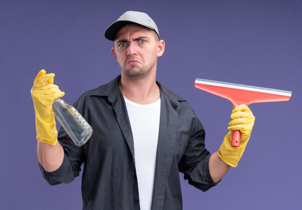 人年轻帅气的清洁工穿着t恤 戴着帽子和手套 拿着拖把头和喷雾瓶隔离在紫色的墙上帽子拖把站着