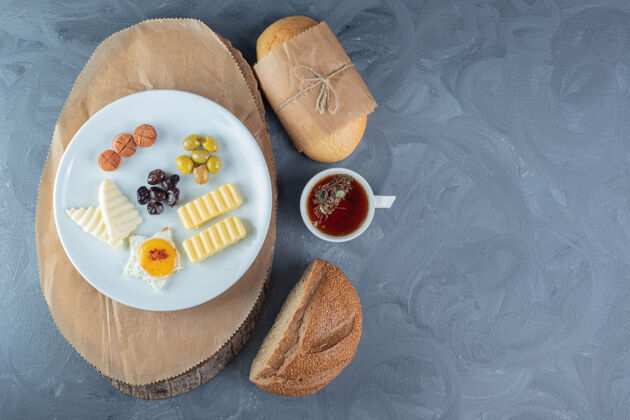 美味在大理石桌上的木板上放着一块面包 一杯茶 一杯果汁 一盘奶酪 鸡蛋 黄油和香肠片切片面包果汁
