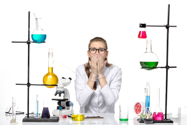表情前视图穿着医疗服的女化学家坐在白色背景上 带着溶液和困惑的表情 化学大流行的冠状病毒化学专业医生
