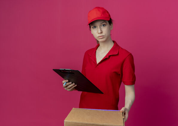 深红色年轻漂亮的送货女孩 穿着红色制服 戴着帽子 手里拿着披萨包和剪贴板 看着被隔离在深红色背景上的相机 还有复印空间红色送货背景