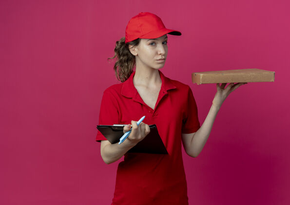 红色自信的年轻漂亮的送货女孩 穿着红色制服 戴着披萨包装的帽子 手里拿着一支笔和剪贴板 背景是深红色 有复印空间制服包装女孩