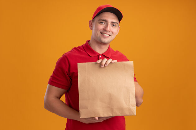 帽子微笑着的年轻送货员穿着制服 戴着帽子 手里拿着隔离在橙色墙上的纸食品包衣服包装站着