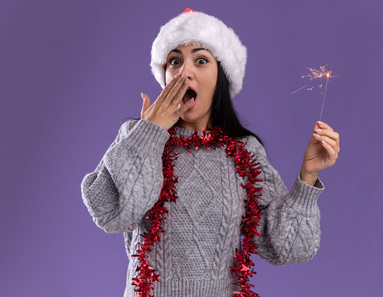 新年关心的年轻白人女孩戴着圣诞帽 脖子上戴着金属丝花环 手里拿着节日火花灯 看着相机 手放在嘴上 在紫色的背景下与世隔绝嘴年轻闪闪发光
