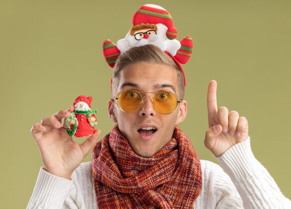 圣诞老人印象深刻的年轻帅哥戴着圣诞老人的头带和围巾看着相机拿着雪人圣诞饰品看着相机指着橄榄绿的背景孤立向上指向装饰品