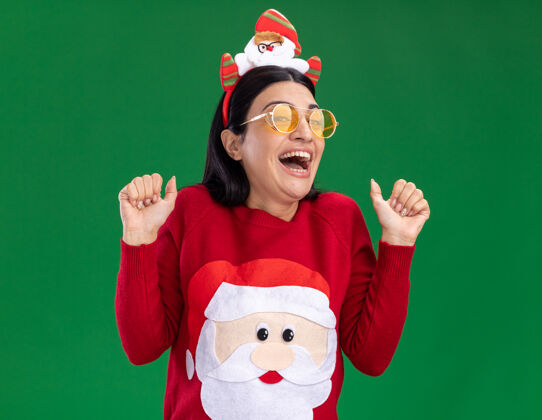 年轻兴奋的年轻白人女孩戴着圣诞老人的头带和毛衣 戴着眼镜 看着相机 在绿色背景上孤立地竖起大拇指圣诞老人眼镜毛衣