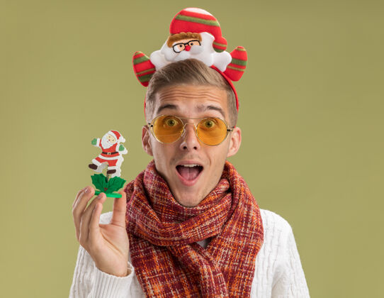 抱印象深刻的年轻帅哥戴着圣诞老人头带和围巾看着相机拿着圣诞老人玩具孤立在橄榄绿的背景圣诞老人绿色头带