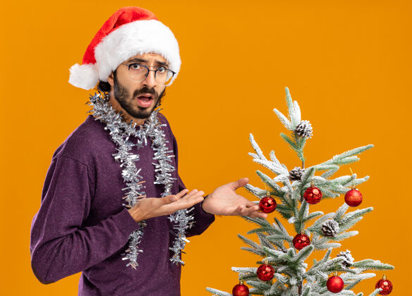 脖子困惑的年轻帅哥站在圣诞树旁 戴着圣诞帽 脖子上戴着花环 指着橘黄色背景上孤立的圣诞树举行圣诞树家伙