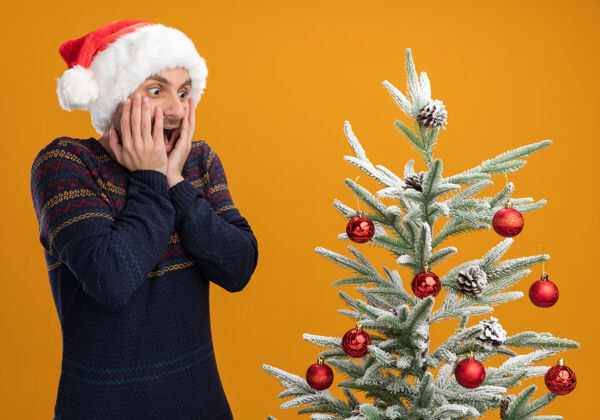 手令人印象深刻的年轻白种人戴着圣诞帽站在装饰圣诞树附近 手放在脸上看着橙色背景上孤立的圣诞树脸圣诞橘子