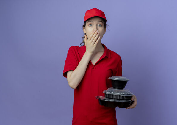 交货令人惊讶的年轻漂亮的送货女孩穿着红色制服 戴着帽子拿着食物容器 手放在嘴上 紫色背景上有复制空间帽子女孩制服