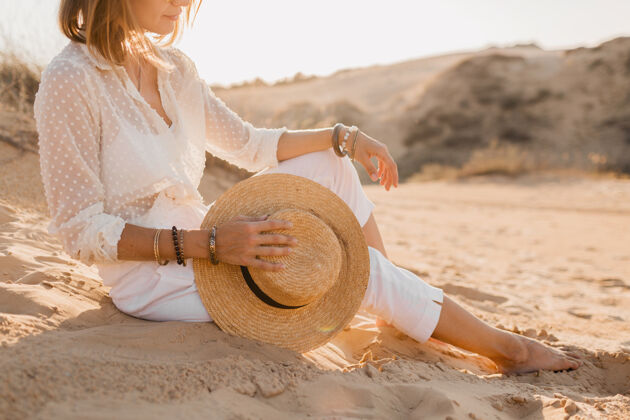 女特写时尚配饰的时尚美女在沙漠沙滩上穿着白色服装手持草帽在日落女人帽子配饰