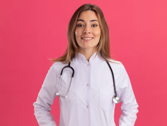 医学微笑着看着前面穿着医用长袍的年轻女医生 听诊器被隔离在粉红色的墙上姿势年轻人医生