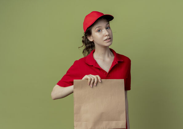 橄榄色令人印象深刻的年轻漂亮的送货女孩在红色制服和帽子举行纸包看着相机隔离在橄榄绿的背景与复印空间帽子背景印象