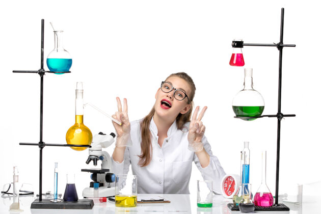 医学前视图：身着医疗服的女化学家坐在桌子前 用白色背景上的解决方案大流行化学冠状病毒V符号烧杯化学
