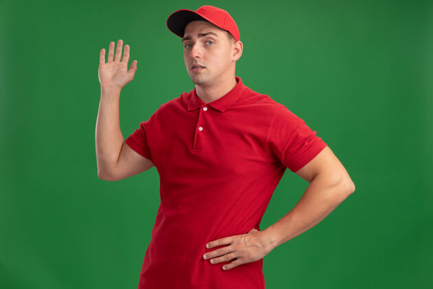 臀部自信的年轻送货员穿着制服 戴着帽子 展示着问候的手势 手放在屁股上 隔离在绿色的墙上交货穿手势
