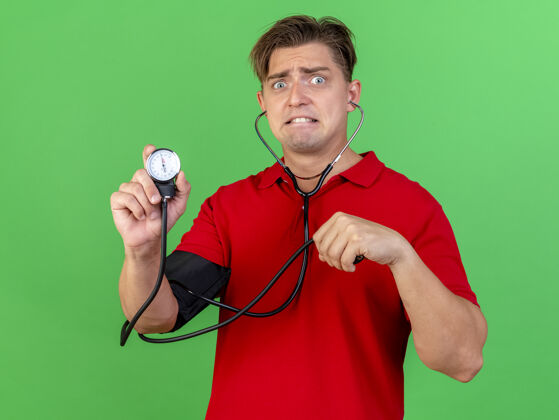 脸焦急的年轻英俊的金发病男子戴着听诊器给自己量血压 拿着血压计看着前面隔离的绿墙上衣服人人