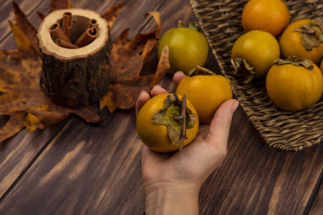 木头女性手拿着新鲜柿子和肉桂棒放在木坛上的顶视图 木桌上放着树叶棒橘子新鲜