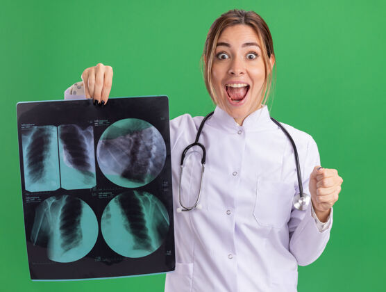 表演兴奋的年轻女医生穿着医用长袍 听诊器拿着x光片 显示“是”的手势 隔离在绿色的墙上医生医疗X光