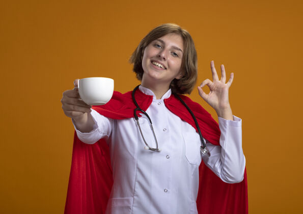 茶身着红色斗篷 面带微笑的金发女超人 身穿医生制服 手持听诊器 朝前端出一杯茶 注视着前方 橙色墙壁上孤立的“一切正常”标志女孩杯子穿着